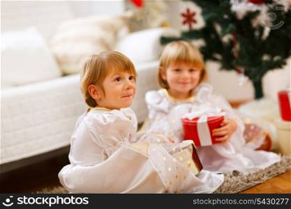 Two twins girls sitting with presents near Christmas tree