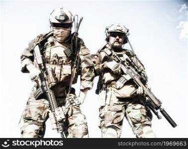 Two U.S. marine riders, military company mercenary in helmets with radio headset, woodland camo battle dress uniform, load carriers, looking down at camera while standing with assault rifles in hands. Army elite soldiers in full tactical ammunition