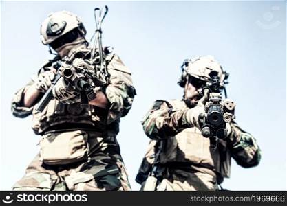 Two U.S. marine riders, military company mercenary in helmets with radio headset, woodland camo battle dress uniform, load carriers, looking down at camera while standing with assault rifles in hands. Army elite soldiers in full tactical ammunition