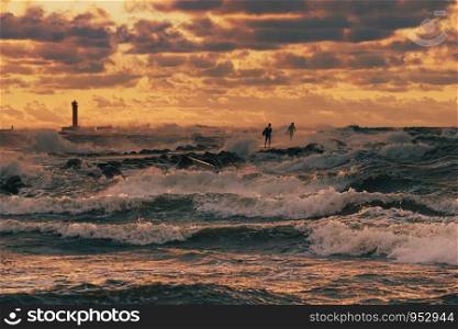 Two unrecognizable kite surfer running around the pier in the stormy sea lit by the sunset light under dramatic orange evening clouds, with a silhouette of lighthouse in the background