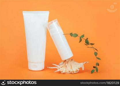 Two white blank cosmetics tube and bottle, sea shell and green eucalyptus branch on orange background. Natural Organic Spa Cosmetic Beauty Concept. Mockup Front view.. Two white blank cosmetics tube and bottle, sea shell and green eucalyptus branch on orange background. Natural Organic Spa Cosmetic Beauty Concept. Mockup Front view
