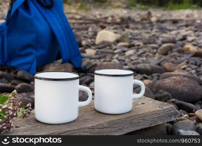 Two white campfire enamel coffee mug mockup with blue backpack. Empty mug mock up for design promotion.
