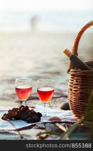 Two wine glasses with red wine standing beach, beside grapes and wicker basket with bottle of wine. Young couple standing in the sea in the background