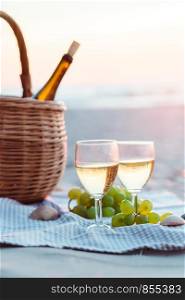 Two wine glasses with white wine standing on sand, on beach, beside grapes and wicker basket with bottle of wine. Sea waves in the background