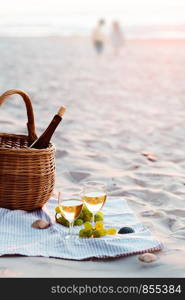 Two wine glasses with white wine standing on sand, on beach, beside grapes and wicker basket with bottle of wine. Young couple standing in the sea in the background