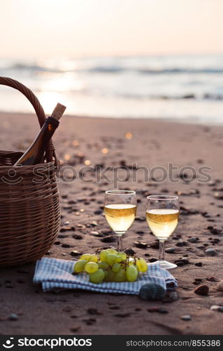 Two wine glasses with white wine standing on sand, on beach, beside grapes and wicker basket with bottle of wine. Sea waves in the background