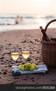 Two wine glasses with white wine standing on sand, on beach, beside grapes and wicker basket with bottle of wine. Young couple standing in the sea in the background
