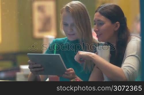 Two women are sitting in cafe and laughing at something they see in tablet PC. Shot is made through cafe show window, rain drops are seen on it.