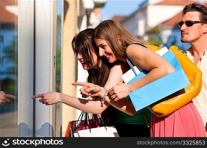 Two women being friends shopping downtown with colorful shopping bags, they are lolling into a glass store door and are amazed; in the background a man is standing