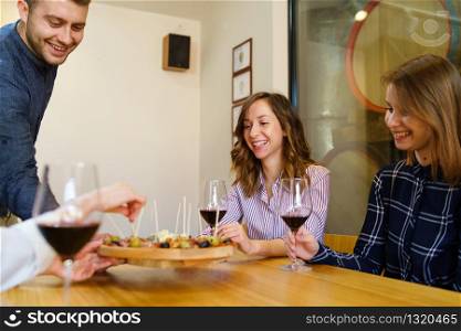 Two women caucasian girls sitting by the table holding glasses of red wine while one young man is serving appetizer friends smiling in day at home or restaurant