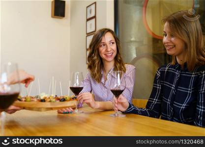 Two women caucasian girls sitting by the table holding glasses of red wine while unknown man is serving appetizer friends smiling in day at home or restaurant