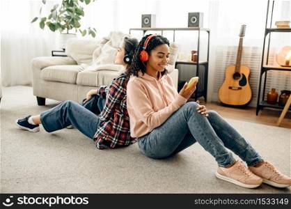 Two women enjoys listening to music sitting back to back on the floor. Pretty girlfriends in earphones relax in the room, sound lovers resting on couch, female friends leisures together. Two women listening to music sitting back to back