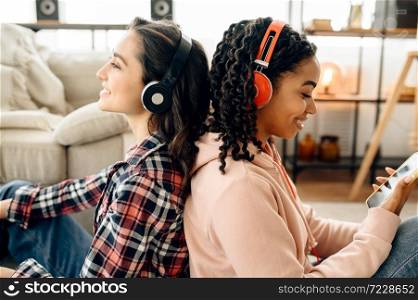 Two women enjoys listening to music sitting back to back on the floor. Pretty girlfriends in earphones relax in the room, sound lovers resting on couch, female friends leisures together. Two women listening to music sitting back to back