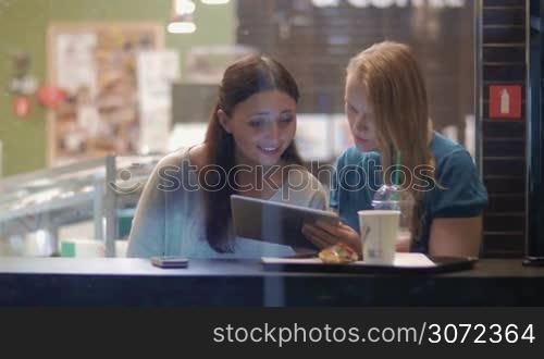 Two women friends meeting in cafe. They having a conversation while looking at pad screen. View through the window, raining outside