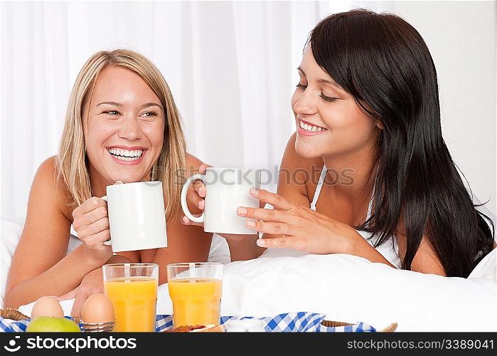 Two women having home made breakfast in white bed, drinking coffee