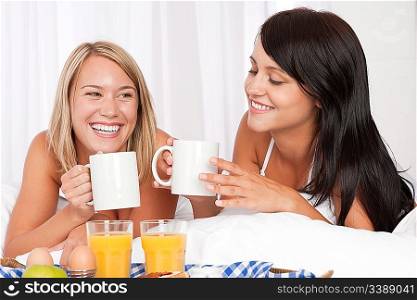 Two women having home made breakfast in white bed, drinking coffee