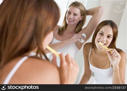 Two women in bathroom brushing teeth applying deodorant and smiling