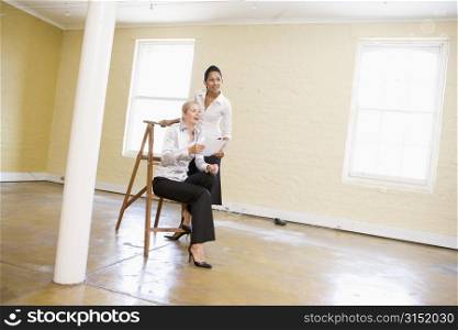 Two women in empty space with ladder holding paper and smiling
