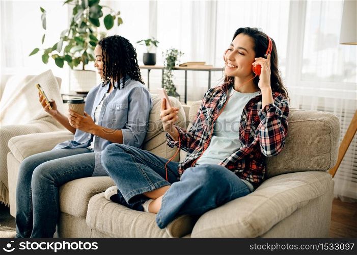 Two women in headphones leisures on sofa. Pretty girlfriends in earphones relax in the room, sound lovers resting on couch, female person using mobile phone. Two women in headphones leisures on sofa