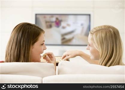 Two women in living room watching television eating chocolates smiling