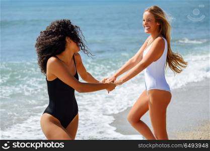 Two women in swimsuit having fun on the beach. Two young women with beautiful bodies in swimwear having fun with their hands caught on the beach. Funny caucasian and arabic females wearing black and white swimsuits