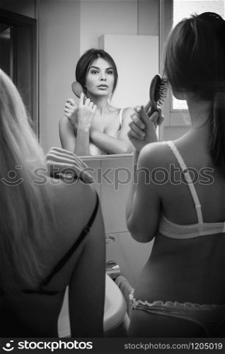 Two women in underwear brushing their hair in the mirror of their bathroom.