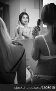 Two women in underwear brushing their hair in the mirror of their bathroom.