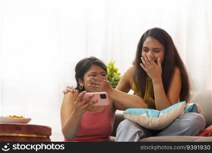 Two  women laugh to≥ther, as they watch a video on mobi≤pho≠whi≤sitting in living room