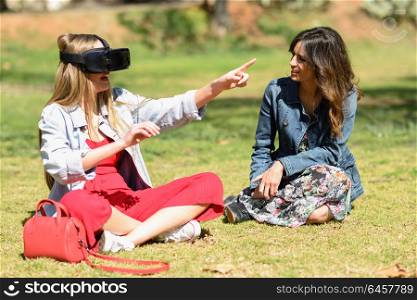 Two women looking in VR glasses and gesturing with his hands outdoors in urban park