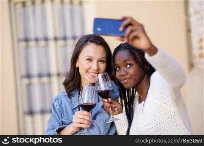 Two women making a selfie with a smartphone while having a glass of wine on the terrace of a bar. Multiethnic women.. Two women making a selfie with a smartphone while having a glass of wine.