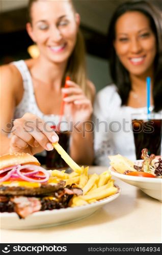 Two women - one is African American - eating hamburger and drinking soda in a fast food diner; focus on the meal