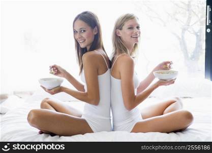 Two women sitting on bed eating cereal smiling