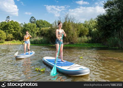 Two women stand up paddleboarding on lake. Young women doing watersport on lake. Female tourists during summer vacation.