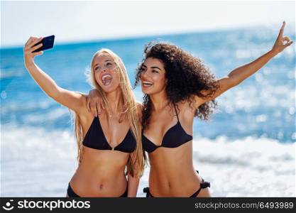 Two women taking selfie photograph with smartphone in the beach. Two young women taking selfie photograph with smart phone in swimwear on a tropical beach. Funny caucasian and arabic females wearing black bikini.