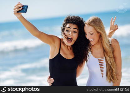 Two women taking selfie photograph with smartphone in the beach. Two young women taking selfie photograph with smart phone in swimsuits on a tropical beach.