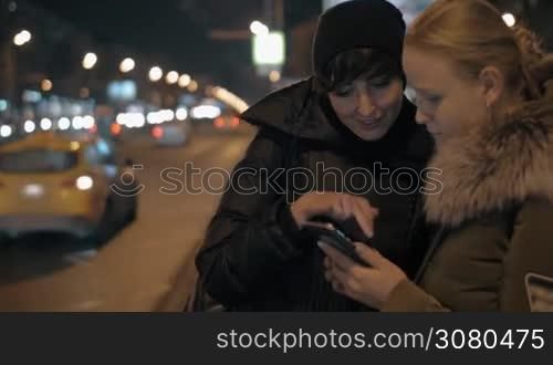 Two women waiting for a bus at public transport stop and surfing internet on smart phone