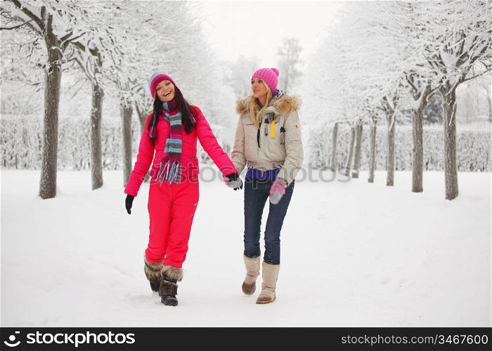 two women walk by winter alley snow trees on background