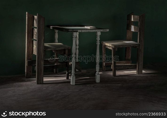 Two wooden chairs and old wooden white table standing front of green cement wall at an empty room through which a ray of light passes. Dark tone, Space for text, Selective focus.
