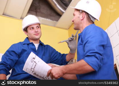 two workers dressed in blue jumpsuits in a house under construction, one of them is taking a drawing