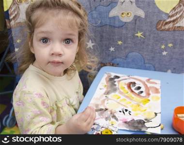 Two-year-old girl is painting at table in children&#39;s room. Landscape page with scribbles and smileys. girl surprised looks into frame.