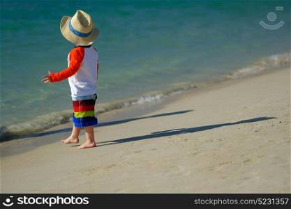Two year old toddler boy in sun hat walking on beach