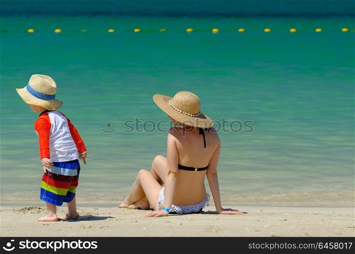 Two year old toddler boy in sun hat with mother on beach