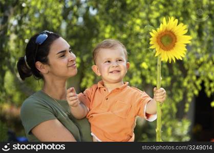 Two years old baby boy and mother enjoy the summer together, playing with a sunflower in the garden. Outdoor, sunlight.