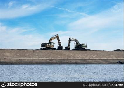 two yellow heavy bulldozer on sand dike with water in front and blue sky and white clouds as background