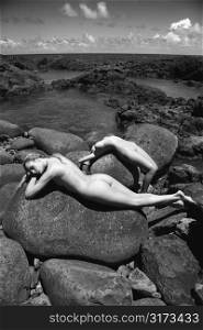Two young adult nude Caucasian women lying on boulders on rocky Maui coast.