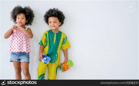 Two young African little boy and girl stand in front of white wall and express different action and they look relax and fun with boy hold some toys in his hand.