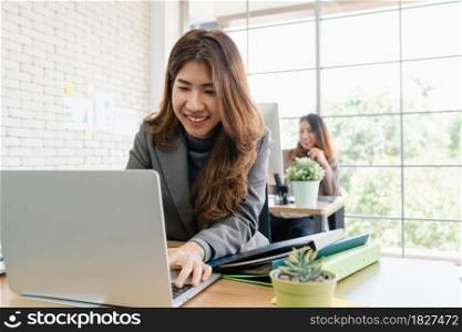 Two young Asian businesswomen working together in office at small business sitting reading a report or paperwork with pleased smiles and successful teamwork. Business, technology and office concept.