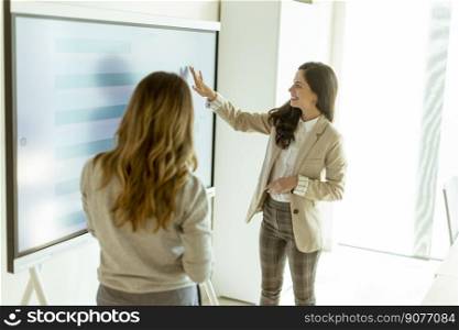 Two young business women discussing financial results on the big wall screen in the office