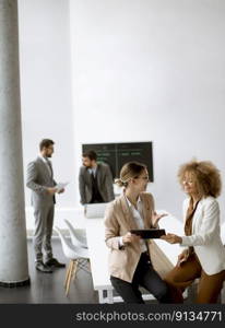 Two young businesswomen discussing with digital tablet in the office with young people works behind them