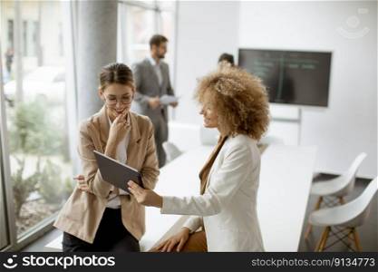 Two young businesswomen discussing with digital tablet in the office with young people works behind them